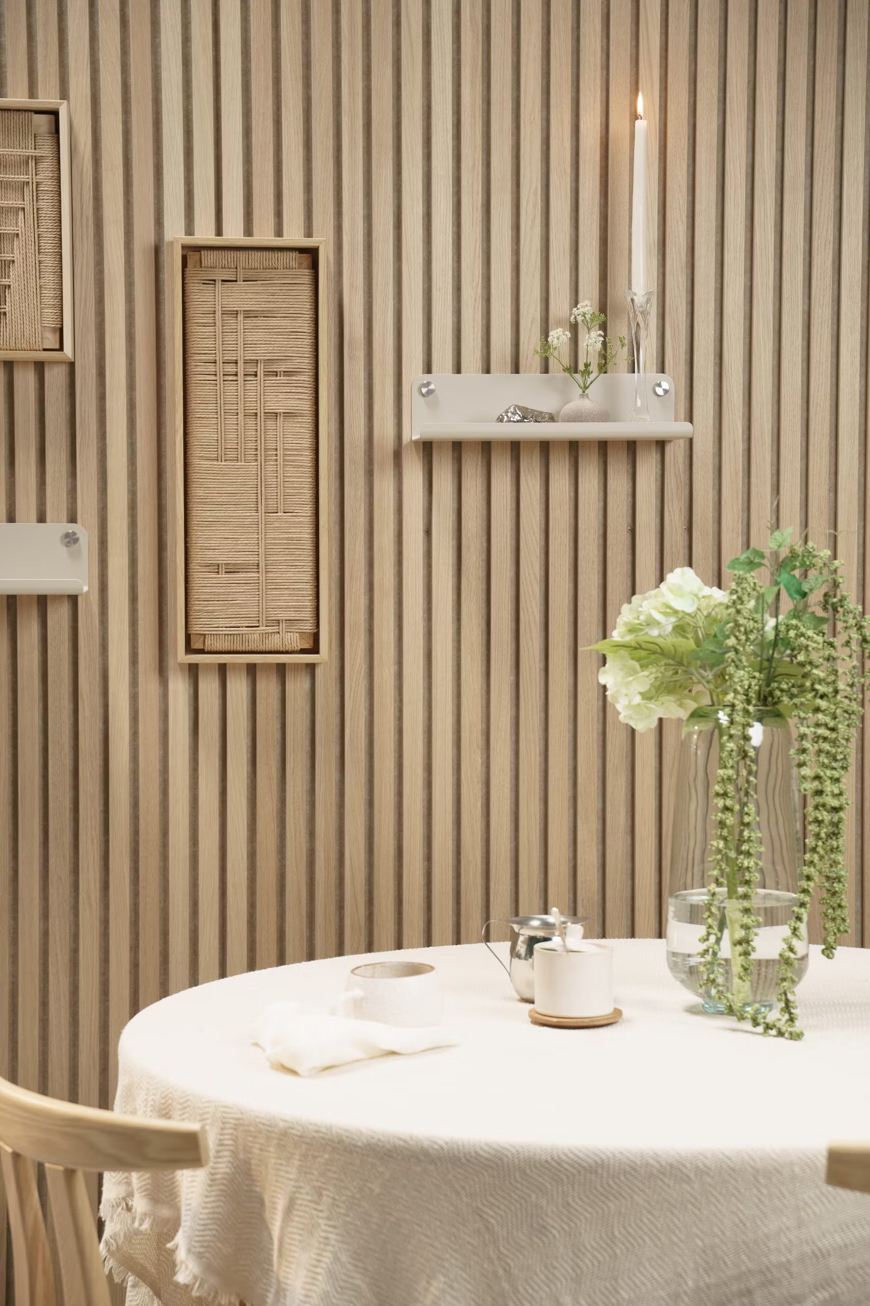 Une table ronde avec une nappe blanche, un vase à fleurs, une bougie et un décor placé devant un mur de lattes acoustiques Akupanel en bois clair avec des accents tissés et des étagères.