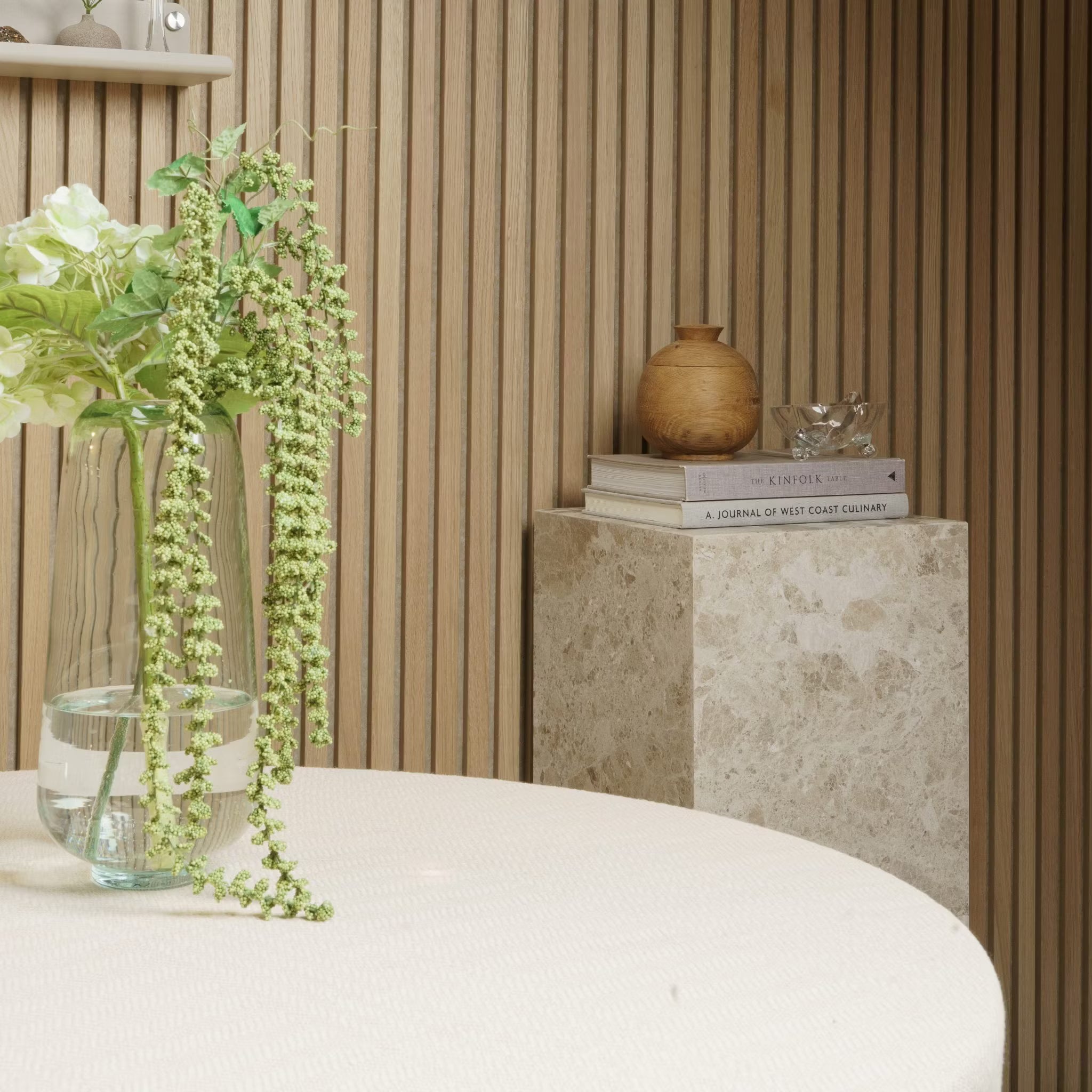 Un vase avec des fleurs est posé sur une table ronde ; des livres et des poteries reposent sur un piédestal en marbre contre un mur en panneaux de bois acoustique Akupanel.
