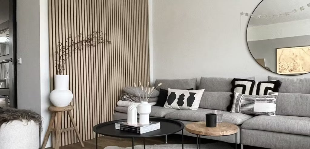 Salon moderne composé dun canapé gris, de coussins abstraits, de tables basses noires, de vases décoratifs et dun mur de lattes acoustiques en bois Akupanel avec un miroir rond.