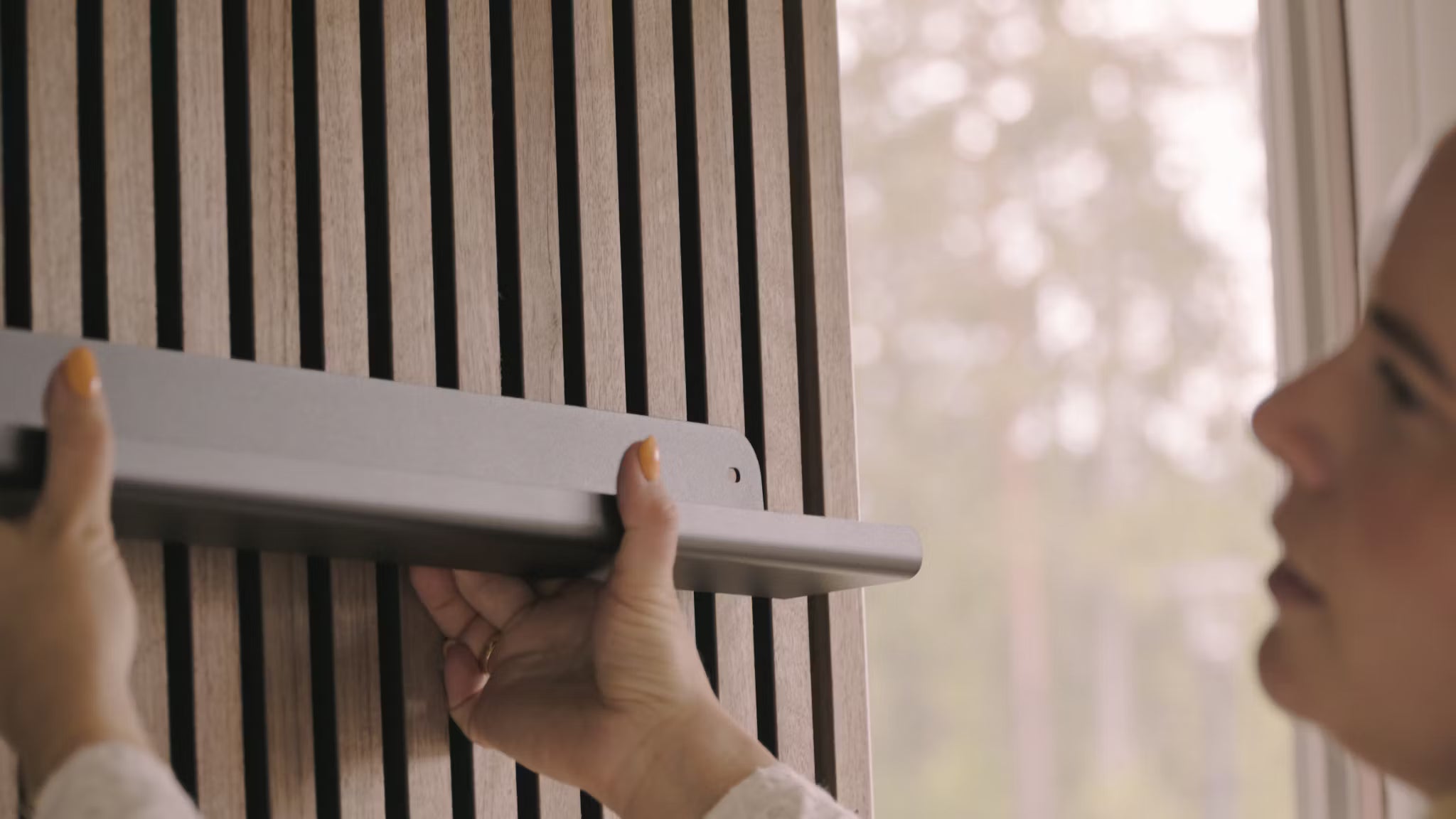 Une personne installe une étagère grise sur un mur de lattes de bois acoustique Nordic Akupanel à côté dune fenêtre donnant sur des arbres.