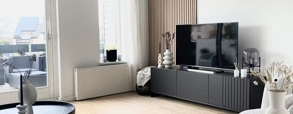 Salon moderne doté dun meuble TV noir, dun téléviseur à écran plat, de vases décoratifs et de grandes fenêtres avec lumière naturelle donnant sur le patio ; Akupanel sur un mur.