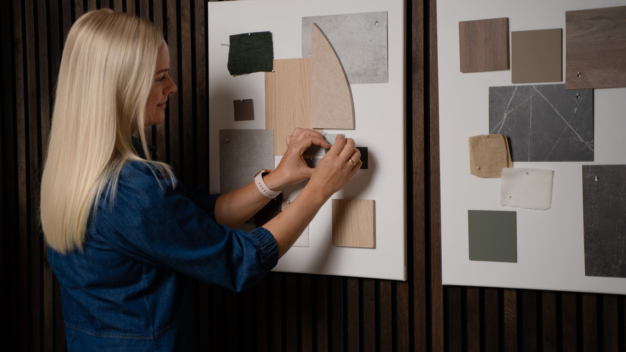 Une femme dispose des échantillons de tissus et de couleurs sur un tableau dhumeur, avec un autre tableau dinspiration à côté delle, près de panneaux acoustiques en bois de la collection Akupanel de WoodUpp.