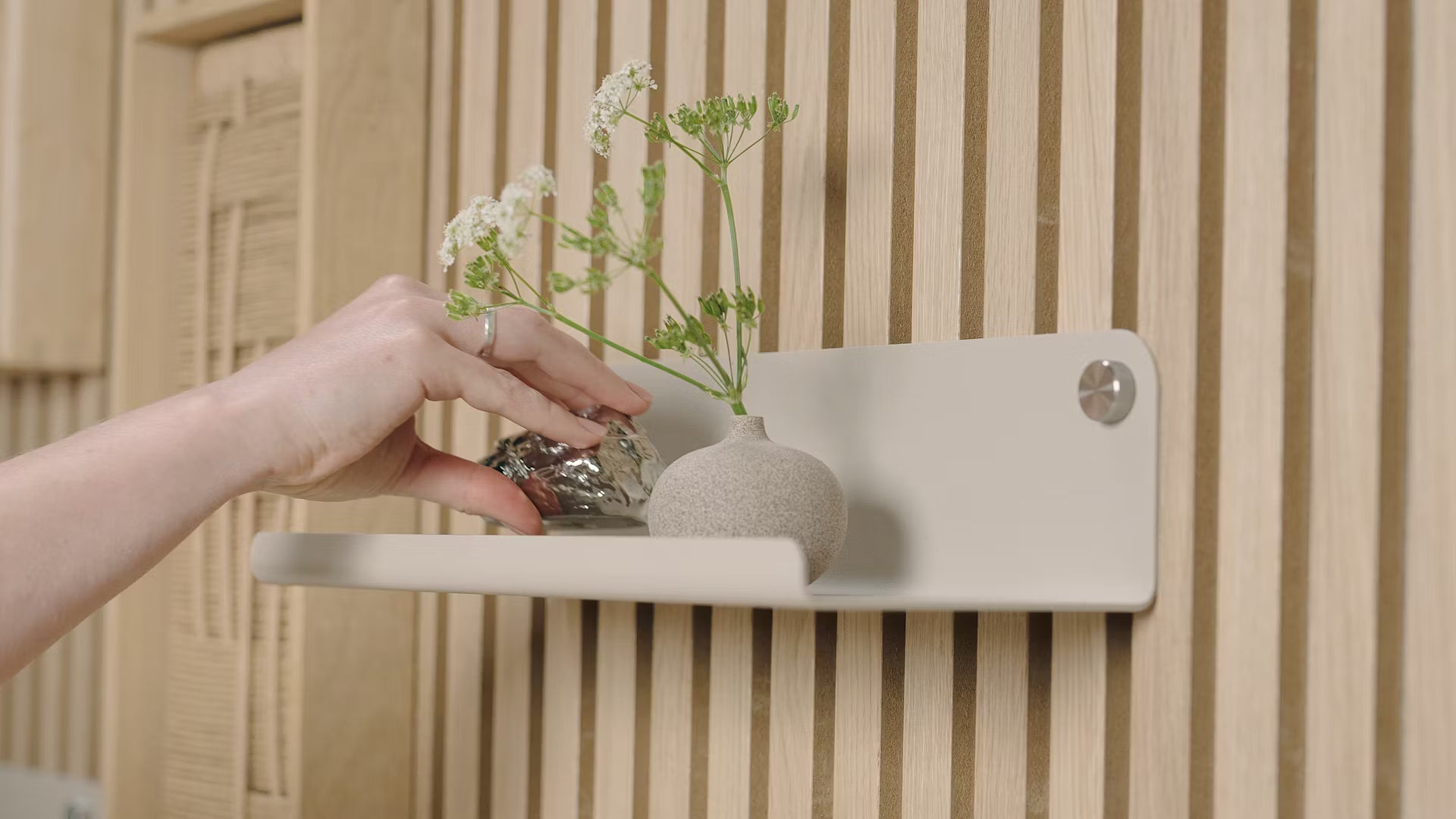 Une main pose un petit objet décoratif à côté dun vase de fleurs blanches sur une étagère minimaliste placée contre un panneau mural en bois acoustique nordique texturé.