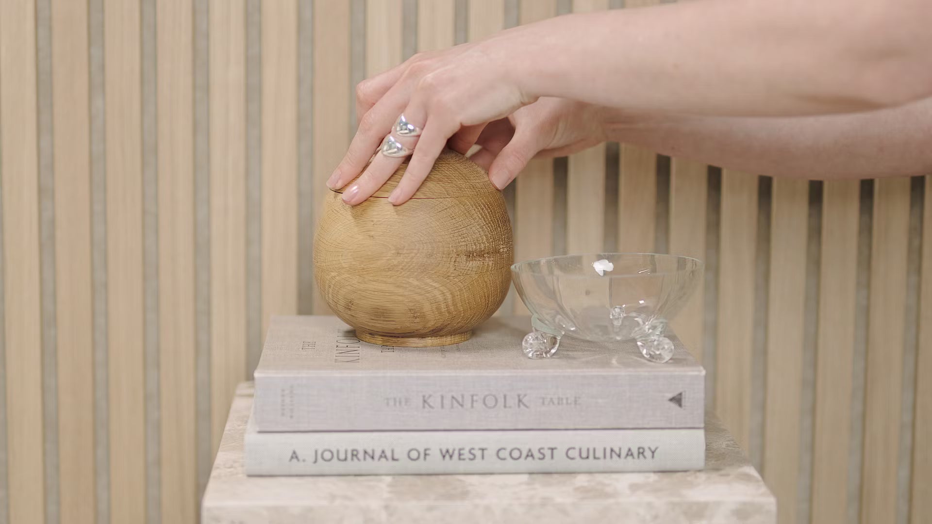 Une main touche une sphère en bois placée sur des livres empilés, dont Kinfolk Table et Journal of West Coast Culinary, à côté dun bol en verre sur un panneau acoustique en lattes de bois.