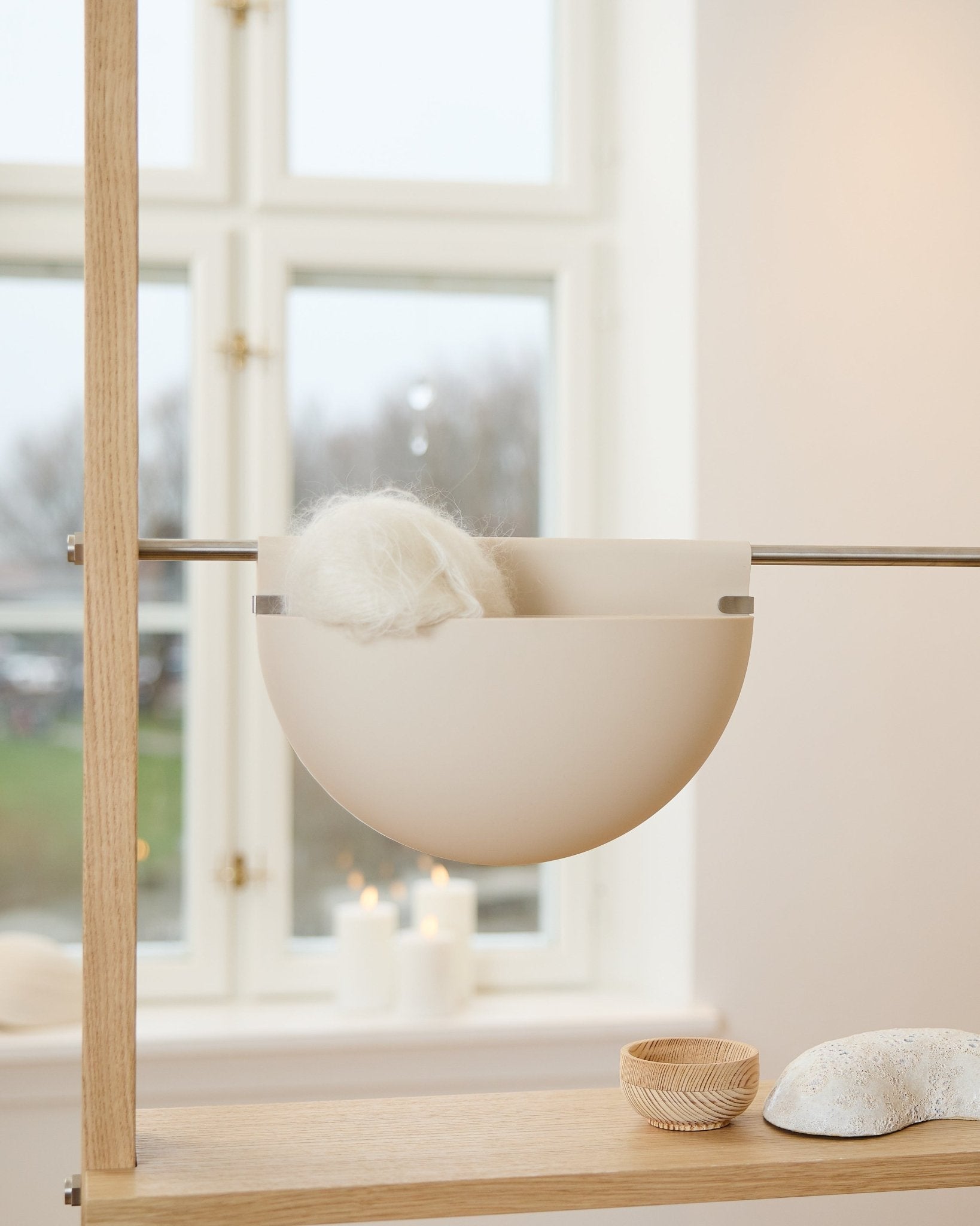 Un bol blanc avec un objet duveteux repose sur une étagère en bois près de bougies, d’objets déco et d’un accessoire acoustique WoodUpp Gro Soft Pocket BM2 en beige clair, près de la fenêtre.