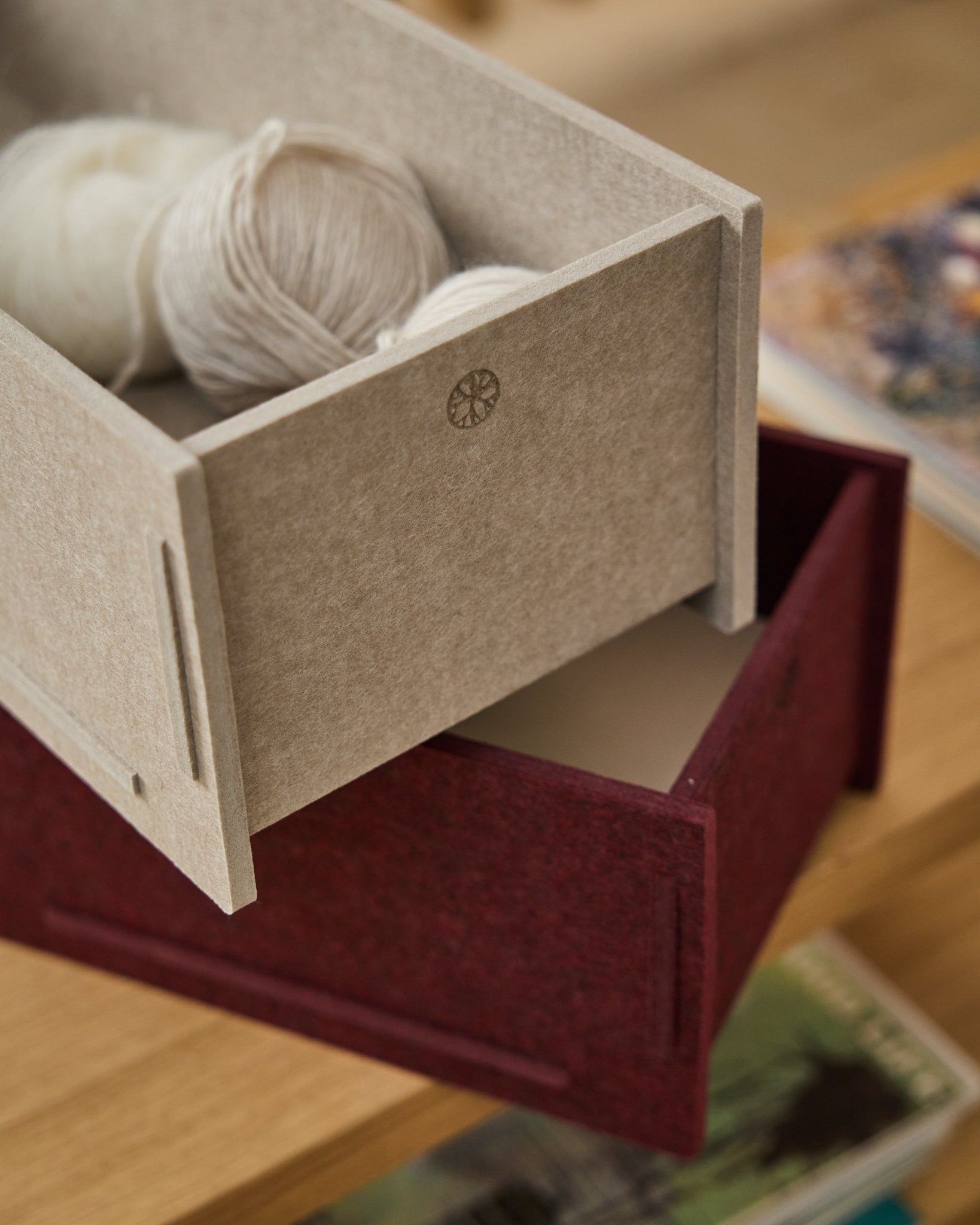 Une boîte de rangement en feutre PET beige, remplie de pelotes de laine, est posée sur une WoodUpp Vigg Felt Box fermée/BM2 en ox red, sur un sol en bois, avec des livres en arrière-plan.