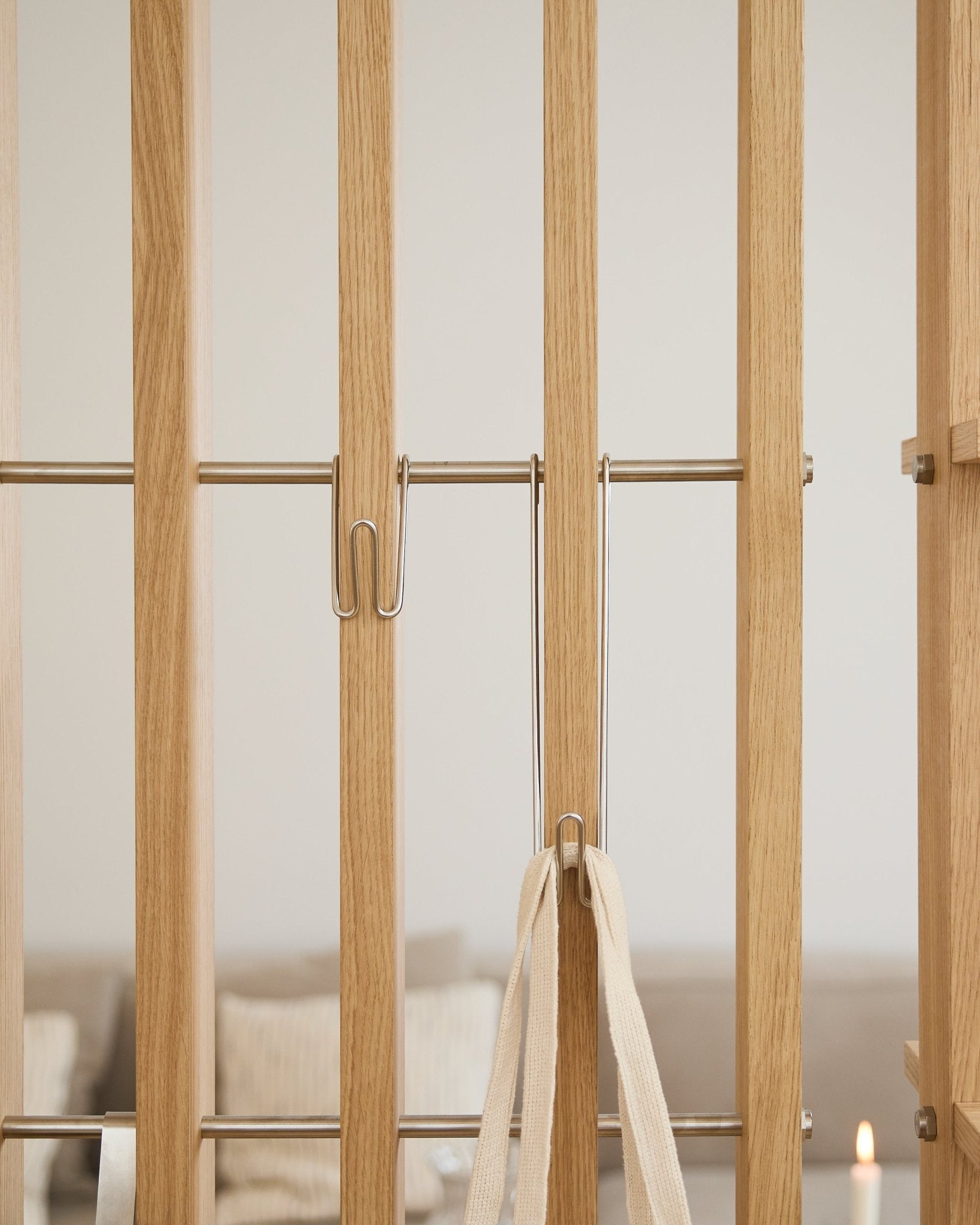 Gros plan sur le WoodUpp Alva | Wire Hook BM1 (acier inoxydable steel, long, lot de 3) fixé à des lattes Panneau acoustique claires avec une sangle blanche ; canapé et bougie flous en arrière-plan.