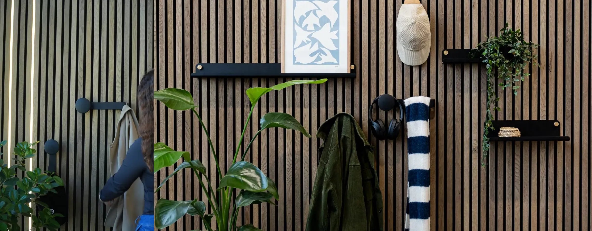 Un homme vêtu dune veste bleue passe devant un mur de lattes en bois acoustique Akupanel nordique où sont intégrés des plantes, des étagères, des patères, des écouteurs et une serviette rayée enroulée.