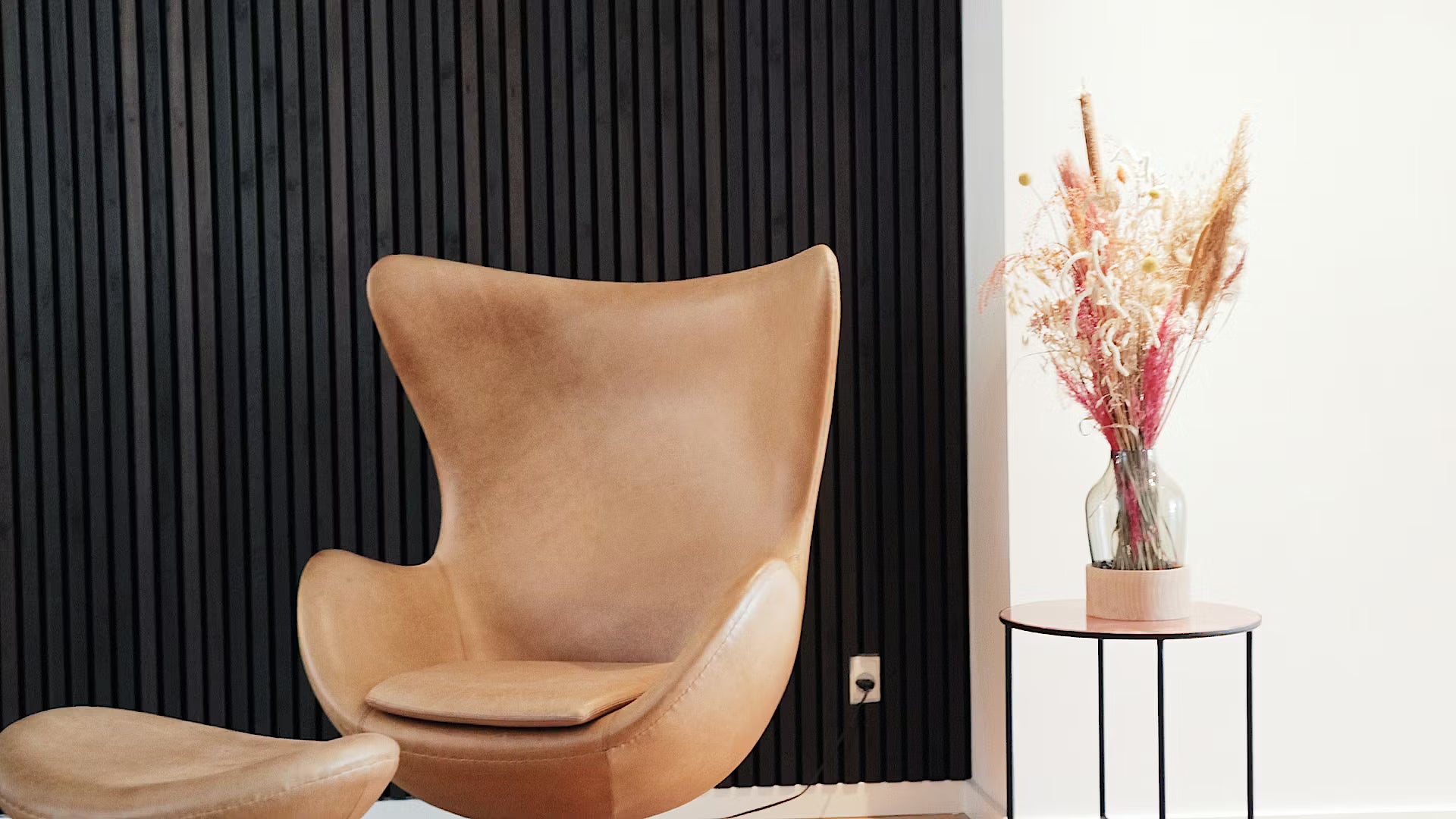 Fauteuil et ottoman en cuir fauve à côté dune petite table avec des fleurs séchées, sur un fond blanc et un panneau de lattes en bois acoustique noir.