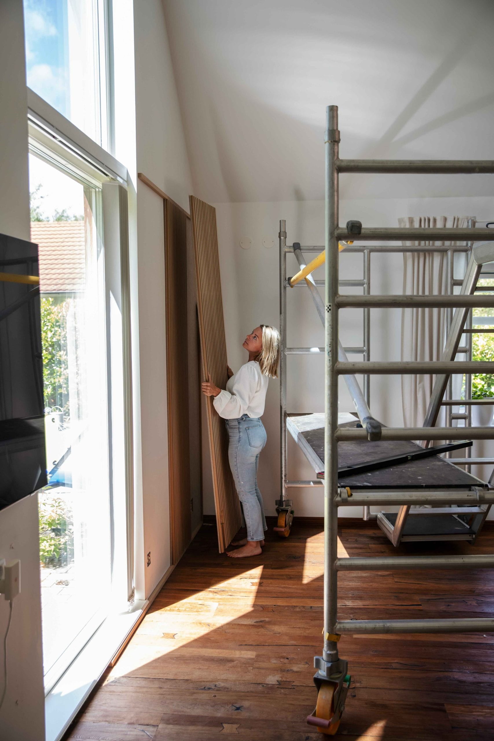 Une femme se tient dans une pièce éclairée par le soleil, dotée de grandes fenêtres et déchafaudages, et tient un panneau de bois acoustique Akupanel posé sur un sol en bois poli.