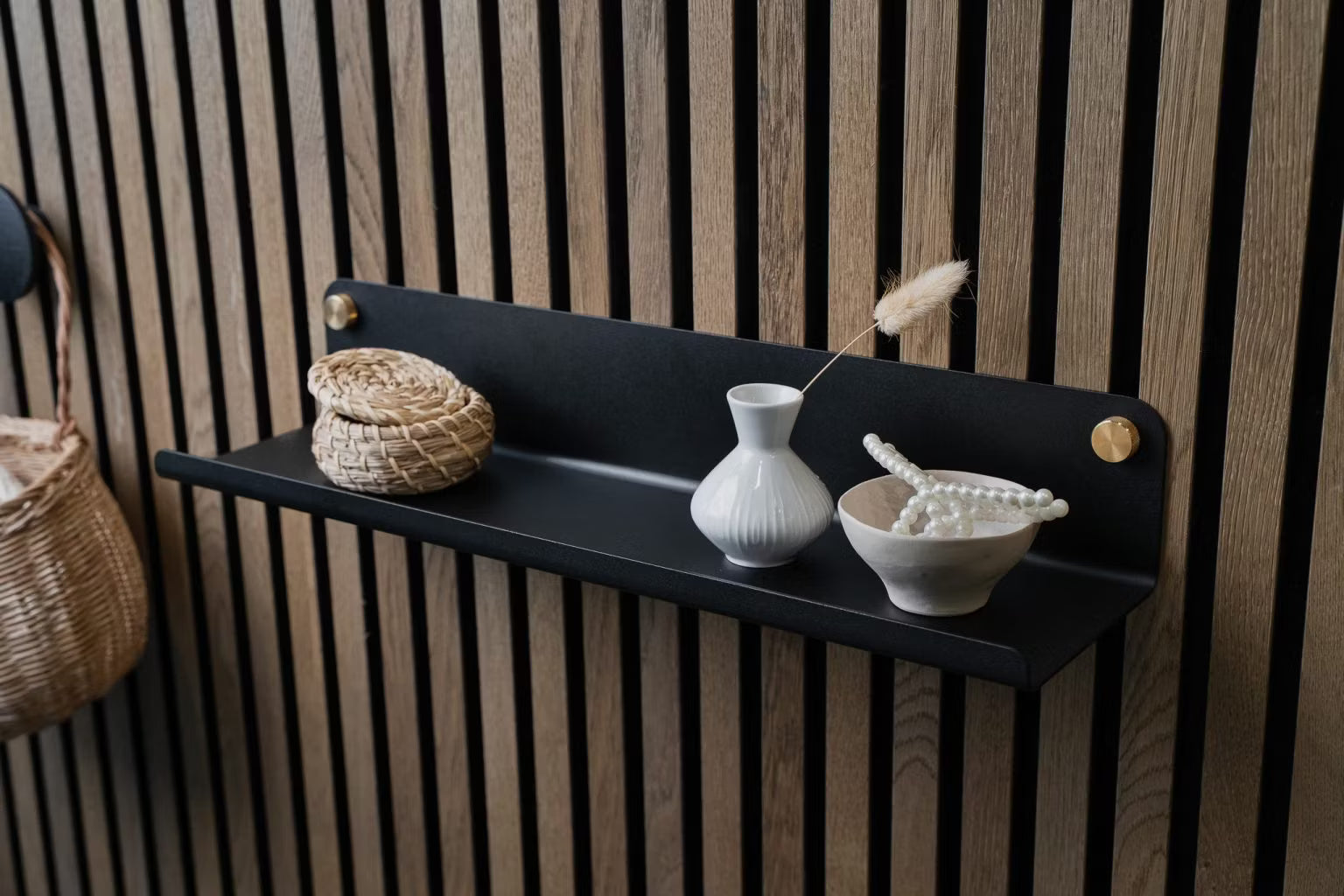 Une étagère murale noire avec un petit panier en osier, un vase blanc et un bol de perles en céramique montés sur un panneau mural acoustique en bois nordique.